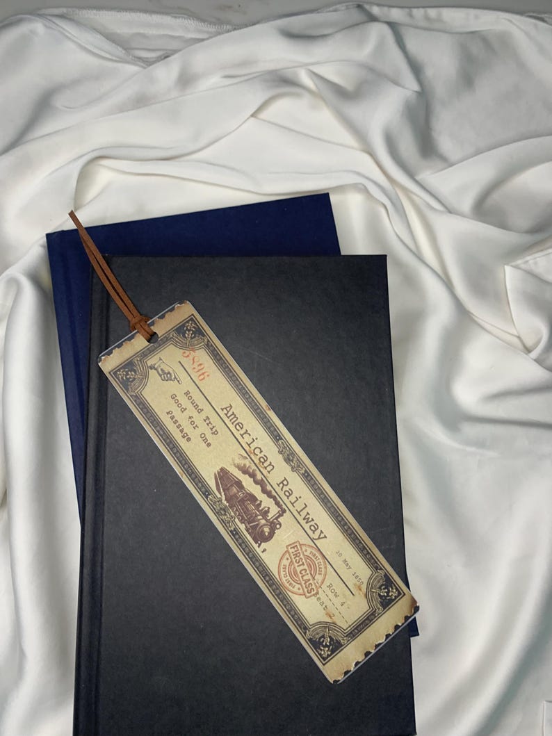 Op de afbeelding: Een vintage-stijl bladwijzer met de tekst "American Railway" en een treinillustratie. De bladwijzer heeft een leren koord en ligt op een zwart boek met een blauw boek erachter. De achtergrond is wit.