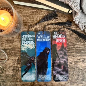 May include: Three rectangular bookmarks featuring artwork from Brandon Sanderson's Mistborn series. Each bookmark displays a hooded figure and the titles The Final Empire, The Well of Ascension, and The Hero of Ages, finished with black tassels.