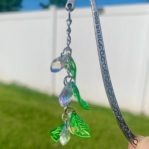 May include: A silver metal bookmark with a decorative floral design. The bookmark features a chain with iridescent crystal-like beads and green leaf accents, creating a whimsical and elegant look. The background is a blurred outdoor scene.
