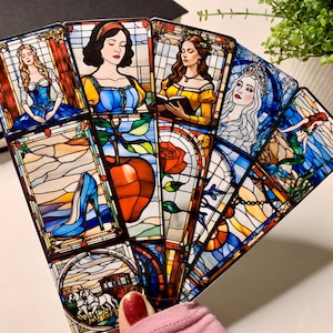May include: A collection of stained-glass style art cards featuring fairytale characters. Each card depicts a different princess, with vibrant colors and intricate details. The cards are held in a hand with a pink sleeve.