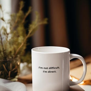 May include: White ceramic mug with the text "I'm not difficult. I'm direct." printed on the front. The mug is sitting on a stack of books.