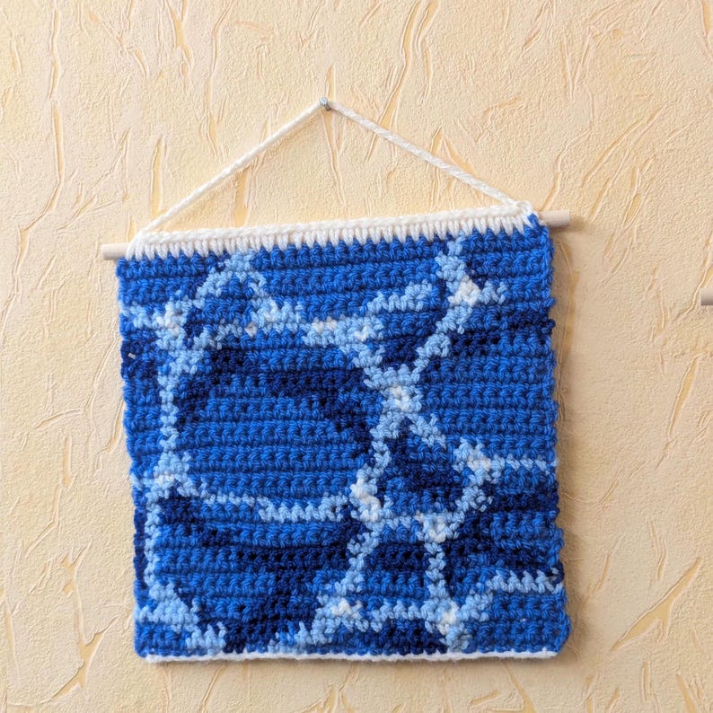 May include: A blue and white crocheted wall hanging with a geometric pattern. The hanging is attached to a wooden dowel rod with white string.