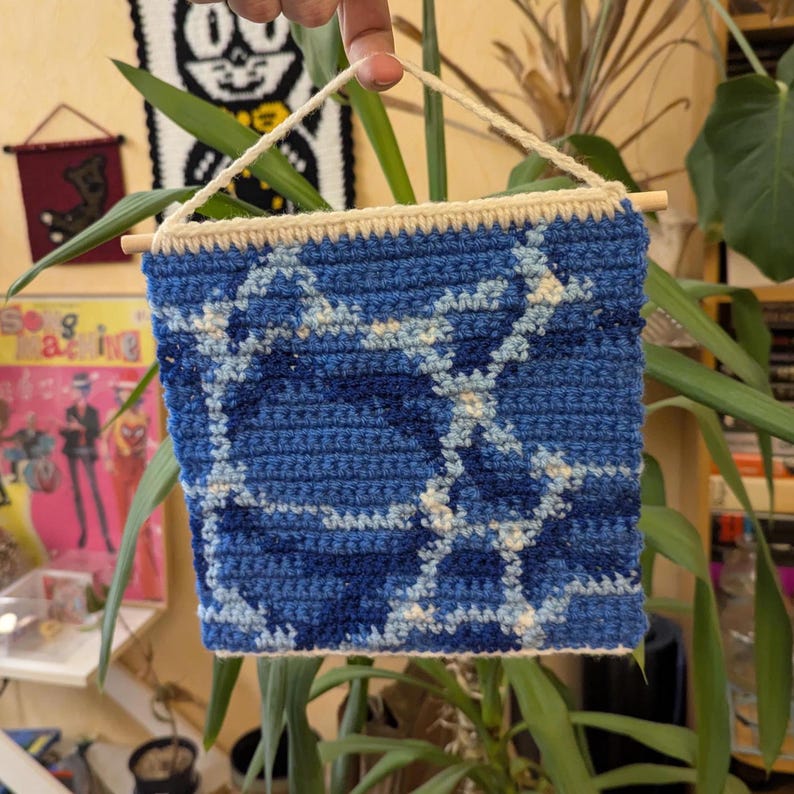 May include: A blue and white crocheted wall hanging with a geometric pattern. The hanging is attached to a wooden dowel rod with a white string.