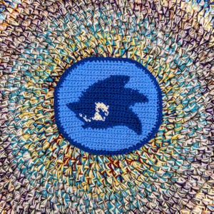 May include: A round, blue crocheted coaster with a dark blue shark silhouette inside a lighter blue circle. The coaster is set on a multicolored, textured surface with shades of blue, yellow, and purple.