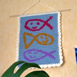 May include: A blue crocheted wall hanging with a white border. The hanging features three colorful fish designs in pink, yellow, and purple.