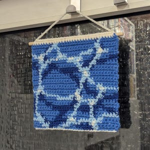 May include: A blue and white crocheted wall hanging with a geometric pattern. The hanging is made with different shades of blue yarn and white yarn.