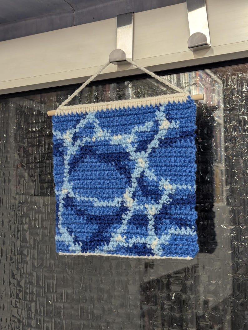 May include: A blue and white crocheted wall hanging with a geometric pattern. The hanging is made with different shades of blue yarn and white yarn.