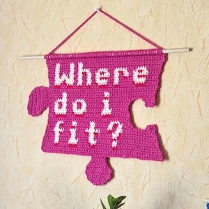 Puede incluir: Colgante de pared de pieza de rompecabezas de crochet rosa con el texto "Where do I fit?" en hilo blanco.