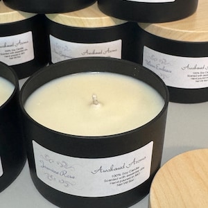 May include: A close-up of a black candle jar with a wooden lid. The candle is labeled "Awakened Aroma" and "Jasmine Rose". The label also states that the candle is 100% soy, scented with essential oils, hand-poured in Missouri, and has a net weight of 6 ounces.