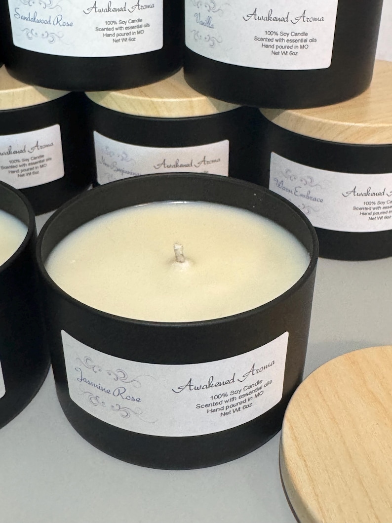 May include: A close-up of a black candle jar with a wooden lid. The candle is labeled "Awakened Aroma" and "Jasmine Rose". The label also states that the candle is 100% soy, scented with essential oils, hand-poured in Missouri, and has a net weight of 6 ounces.