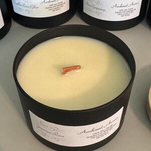 May include: A black candle jar with a wooden lid. The candle is white and has a cinnamon stick on top. The label on the jar reads "Awakened Aromas" and "Jasmine Rose". The candle is 100% soy and hand-poured in the USA.