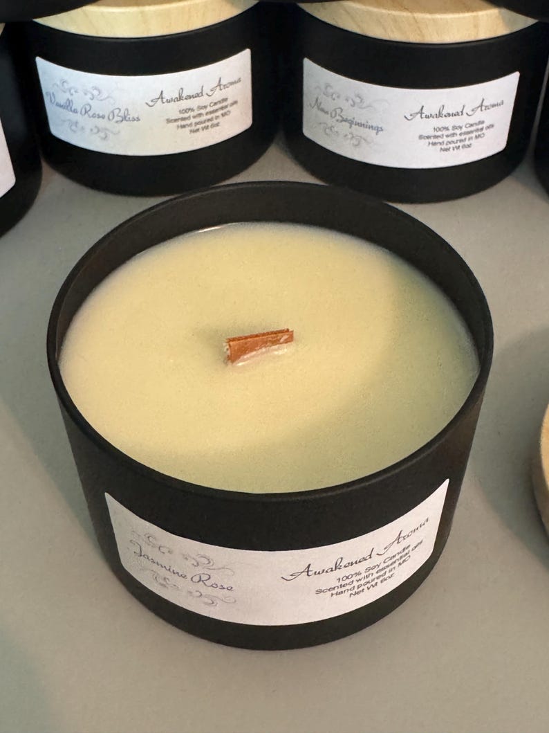 May include: A black candle jar with a wooden lid. The candle is white and has a cinnamon stick on top. The label on the jar reads "Awakened Aromas" and "Jasmine Rose". The candle is 100% soy and hand-poured in the USA.
