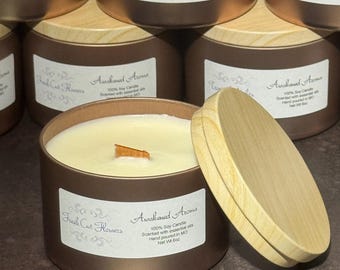 May include: Bronze-colored soy candles with wooden lids. The candles have labels that read "Awakened Aroma" and "Fresh Cut Flowers." The candle wicks are made of wood. The candles are hand-poured and scented with essential oils.