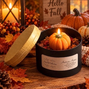 May include: A pumpkin-shaped candle in a black round container with a gold lid. The label reads American Aromas, 100% Soy Candle, Scented with Essential Oils, Hand Poured in the USA, Net Wt 6oz. The candle is lit, revealing a textured orange wax surface.