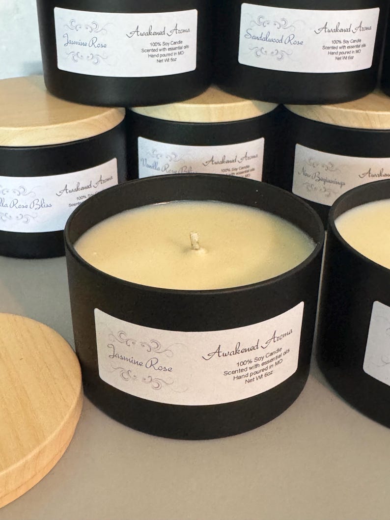 May include: A black candle jar with a wooden lid. The candle is labeled "Jasmine Rose" and "Awakened Aroma" with the text "100% Soy Candle Scented with essential oils Hand poured in MO Net Wt 6 oz".