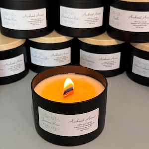 May include: A black soy candle with a wooden lid and a white label that reads "Awakened Aromas" and "Jasmine Rose". The candle is lit and the wax is melting. The candle is 100% soy and scented with essential oils. It is hand-poured in Missouri.
