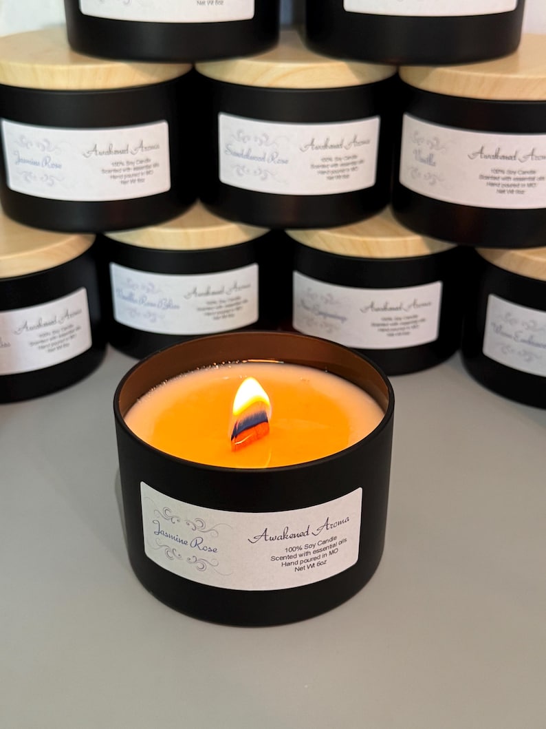 May include: A black soy candle with a wooden lid and a white label that reads "Awakened Aromas" and "Jasmine Rose". The candle is lit and the wax is melting. The candle is 100% soy and scented with essential oils. It is hand-poured in Missouri.