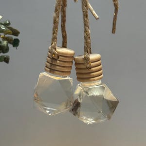 May include: Two clear, faceted glass car air fresheners with wooden caps and braided gold-toned cords. The air fresheners are hanging against a neutral background. The caps are made of stacked wooden rings.
