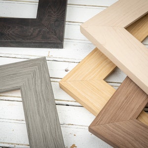 May include: Four different picture frames made of wood, with various wood grain patterns and colors. The frames are arranged in a stack, with the top frame being a light wood color, the second frame being a darker wood color, the third frame being a gray wood color, and the bottom frame being a dark brown wood color.