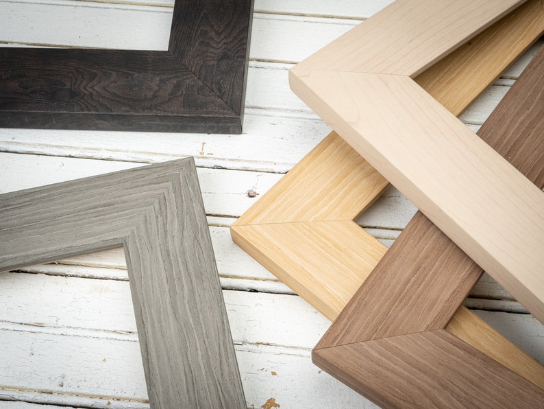 May include: Four different picture frames made of wood, with various wood grain patterns and colors. The frames are arranged in a stack, with the top frame being a light wood color, the second frame being a darker wood color, the third frame being a gray wood color, and the bottom frame being a dark brown wood color.