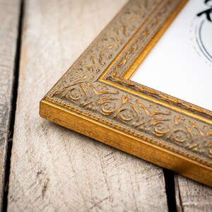 May include: A gold picture frame with an ornate design. The frame is on a light wood surface.