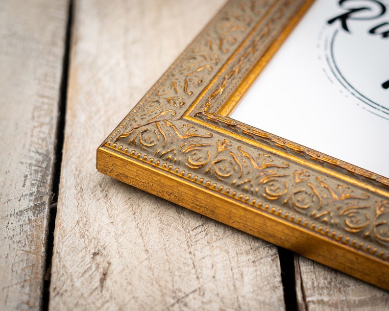 May include: A gold picture frame with an ornate design. The frame is on a light wood surface.