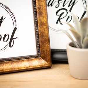 以下が含まれることがあります： 白い背景に黒い円の中に「The Rusty Roof」という文字が書かれた額装されたプリントが2枚。フレームは金色と黒色。フレームの前に小さな鉢植えの植物が置かれている。