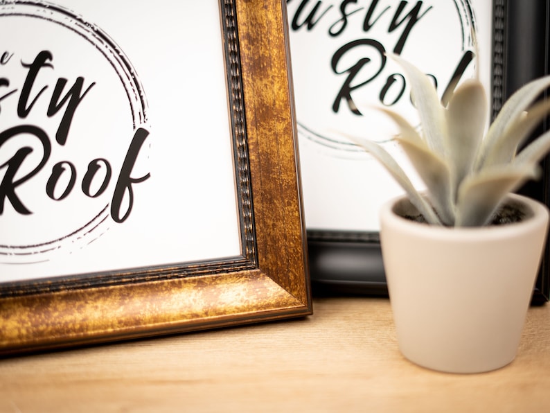 以下が含まれることがあります： 白い背景に黒い円の中に「The Rusty Roof」という文字が書かれた額装されたプリントが2枚。フレームは金色と黒色。フレームの前に小さな鉢植えの植物が置かれている。