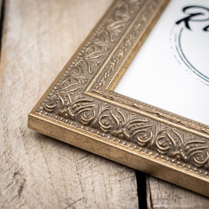May include: A gold picture frame with an ornate design. The frame is on a wooden surface.