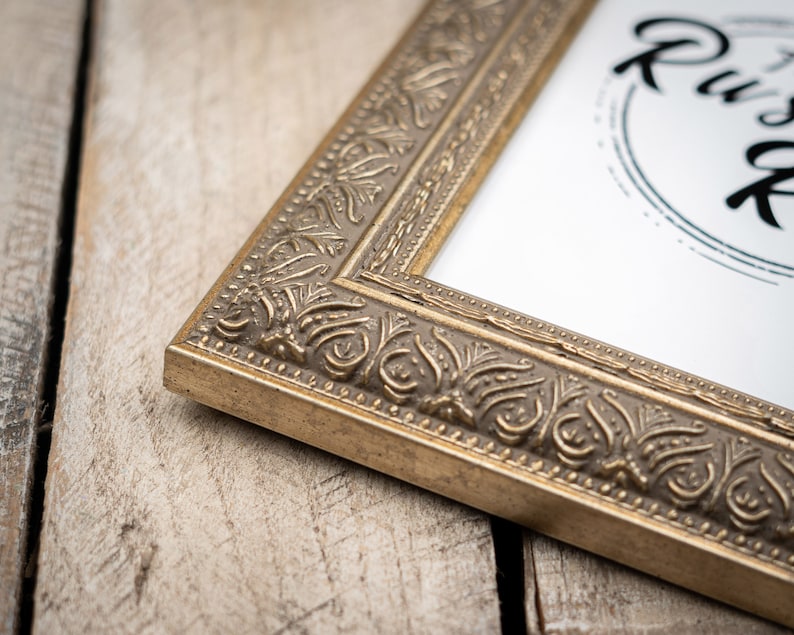 May include: A gold picture frame with an ornate design. The frame is on a wooden surface.