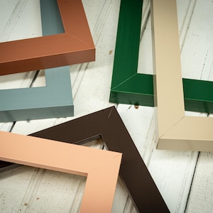 May include: An assortment of colorful picture frame corners in shades of brown, green, gray, and beige. The frames are arranged on a white wooden surface, showcasing their various colors and angles. The frames are made of wood.
