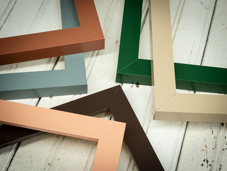 May include: An assortment of colorful picture frame corners in shades of brown, green, gray, and beige. The frames are arranged on a white wooden surface, showcasing their various colors and angles. The frames are made of wood.