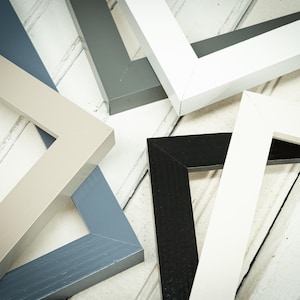 May include: An assortment of wooden picture frames in various colors, including white, black, gray, and blue. The frames are arranged on a light-colored, wooden surface, showcasing their different hues and angles.