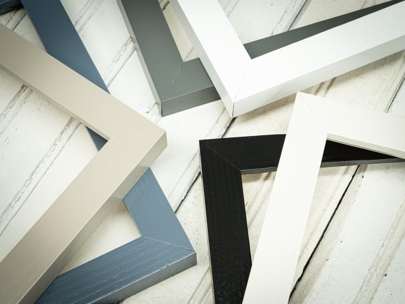 May include: An assortment of wooden picture frames in various colors, including white, black, gray, and blue. The frames are arranged on a light-colored, wooden surface, showcasing their different hues and angles.