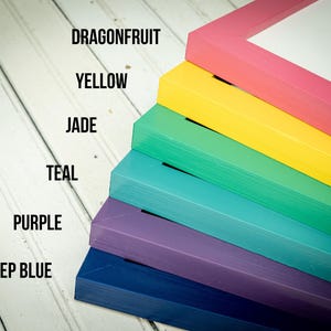 May include: A stack of colorful picture frames in shades of pink, yellow, green, teal, purple, and deep blue. The frames are labeled with color names: Dragonfruit, Yellow, Jade, Teal, Purple, and Deep Blue. The frames are stacked diagonally on a white wooden surface.