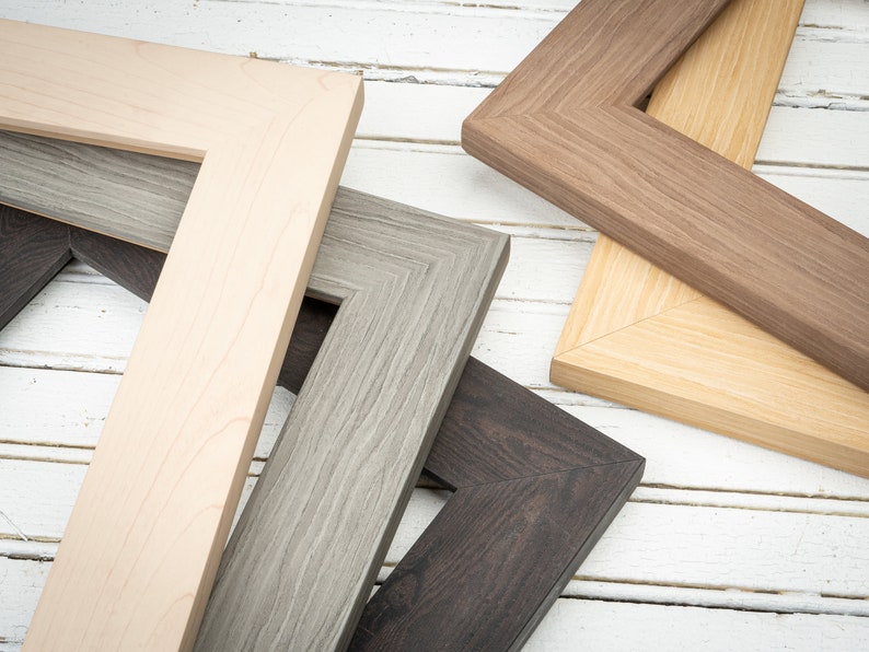 May include: Four different picture frames in various wood tones and finishes. The frames are arranged in a stack, with the top frame being a light brown wood, the second frame being a dark brown wood, the third frame being a gray wood, and the bottom frame being a black wood.