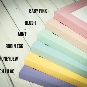 May include: A stack of colorful picture frames in shades of baby pink, blush, mint, robin egg, honeydew, and french lilac. The frame colors are labeled with their names in black text. The frames are angled on a white wooden surface.