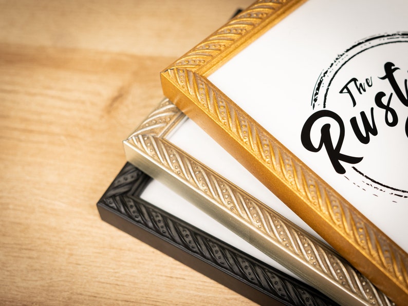 May include: Three picture frames stacked on top of each other. The top frame is gold, the middle frame is silver, and the bottom frame is black. All three frames have a rope-like design around the edges. The top frame has a white paper insert with the text "The Rustic Co." printed on it.