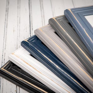 May include: A stack of five rectangular picture frames in black, white, navy blue, light gray, and slate blue. Each frame features a stepped edge profile with visible distressed, worn-away paint along the inner and outer ridges.