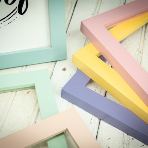 May include: An assortment of colorful picture frames in pastel shades of blue, green, pink, yellow, and purple. The frames are arranged on a white wooden surface, showcasing their various shapes and colors. The frames are ready to be used for displaying photos or artwork.