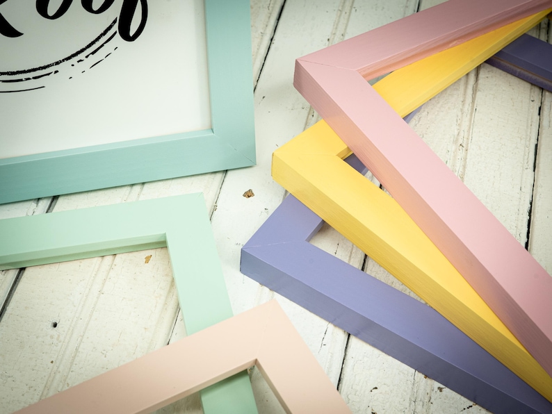 May include: An assortment of colorful picture frames in pastel shades of blue, green, pink, yellow, and purple. The frames are arranged on a white wooden surface, showcasing their various shapes and colors. The frames are ready to be used for displaying photos or artwork.