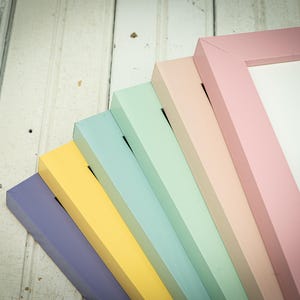 May include: A collection of pastel-colored picture frames in shades of lavender, yellow, light blue, mint green, and pink. The frames are arranged in a fan shape, showcasing their painted edges and the white interior. The background is a white wooden surface.