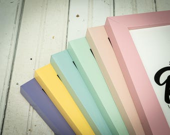 May include: A collection of pastel-colored picture frames in shades of lavender, yellow, light blue, mint green, and pink. The frames are arranged in a fan shape, showcasing their painted edges and the white interior. The background is a white wooden surface.