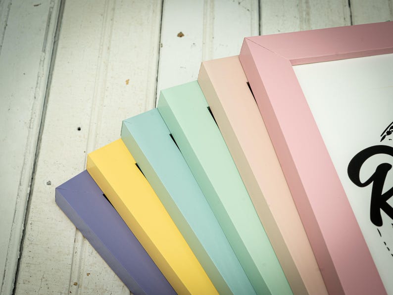 May include: A collection of pastel-colored picture frames in shades of lavender, yellow, light blue, mint green, and pink. The frames are arranged in a fan shape, showcasing their painted edges and corners. The background is a white, weathered wood surface.