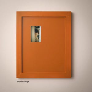 a young boy peeking out from behind a small orange picture frame.