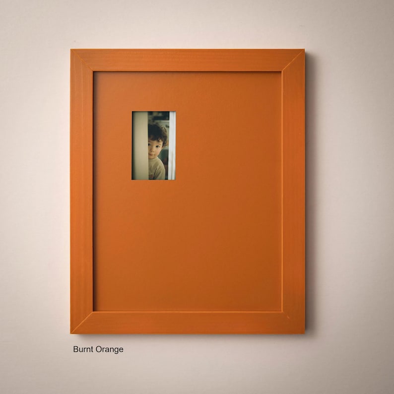 a young boy peeking out from behind a small orange picture frame.