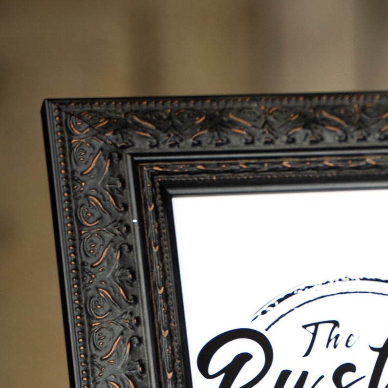May include: Black picture frame with an ornate, detailed design. The frame has a gold-toned finish. The frame is empty and ready to display a photo or artwork.