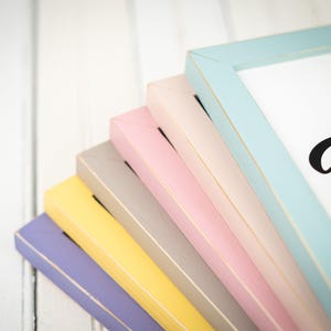 May include: A stack of colorful picture frames in pastel shades of blue, pink, yellow, gray, and purple. The frames are made of wood and have a distressed finish, with a white background.