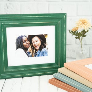 May include: A green picture frame with a white mat holds a photo of two smiling people. Several other frames in shades of peach, beige, and blue are stacked nearby. A small vase with flowers sits to the right.