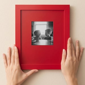 A person's hands are holding a red picture frame with a black and white photograph of two children inside it.
